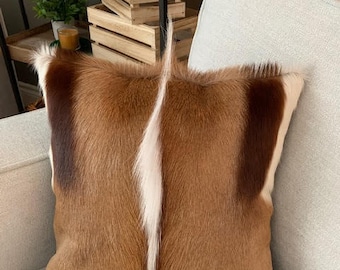 May include: A brown and white cowhide throw pillow with a white stripe down the center. The pillow is on a light beige couch.