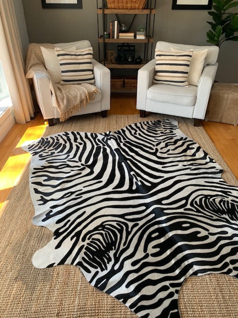 May include: Black and white zebra print cowhide rug with a shaggy texture. The rug is laid out on a woven rug with a light brown color.