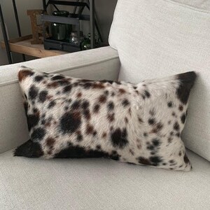 May include: A white and brown cowhide pillow with black spots. The pillow is on a white couch.