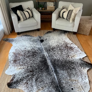 Może przedstawiać: Czarno-biały dywan ze sk&oacute;ry krowiej z naturalnym, cętkowanym wzorem. Dywan leży na tkanym dywanie w salonie z dwoma białymi fotelami i drewnianą p&oacute;łką w tle.