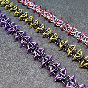 May include: Close-up of three chainmail bracelets in various colors. The bracelets feature intricate interwoven metal rings in purple, gold, and red, with a silver base. The bracelets are displayed on a dark gray surface. The bracelets are a type of jewelry.