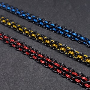 May include: Close-up of three chainmail bracelets in red, gold, and blue. Each bracelet is made of small, interlocking metal rings. The bracelets are displayed on a dark surface.
