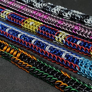 May include: Close-up of several chainmail bracelets in various colors, including black, silver, pink, red, blue, gold, and green. The bracelets are made of small, interlocking metal rings, creating a textured surface. The bracelets are displayed at an angle.