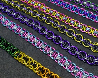 May include: Close-up of several colorful chainmail bracelets. Each bracelet is made of small, interlocking metal rings in various colors, including purple, green, pink, yellow, and black. The bracelets are arranged diagonally against a dark background.