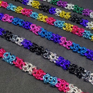 May include: Rows of colorful chainmail jewelry in various colors, including pink, blue, silver, black, yellow, and green. The chainmail is made of small, interlocking metal rings, creating a textured surface. The jewelry is displayed on a dark surface.
