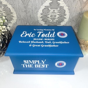 May include: A rectangular blue wooden memorial box featuring white text that reads In Loving Memory Of Eric Todd 08.11.1946 - 08.08.1925 Beloved Husband, Dad, Grandfather & Great Grandfather, with the Rangers Football Club logo and Simply The Best.