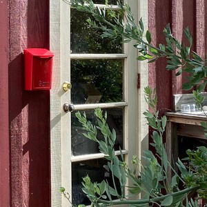 Puede incluir: Un buz&oacute;n rojo con la palabra "Barn" est&aacute; fijado a una pared de madera roja. Una puerta blanca con paneles de vidrio est&aacute; a la derecha del buz&oacute;n. Hay plantas verdes frondosas frente a la puerta.