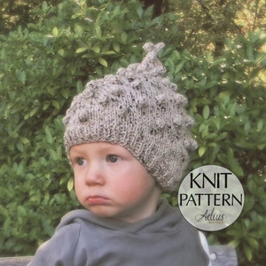 Puede incluir: Un niño con una sudadera con capucha gris y un gorro de punto beige con la parte superior puntiaguda y un diseño texturizado. El gorro tiene una etiqueta "KNIT PATTERN" con el texto "Aelius Boutique" debajo.