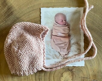 May include: A pink knitted baby bonnet with a tie. The bonnet is made with a soft, textured yarn. The bonnet is lying on a wooden surface.
