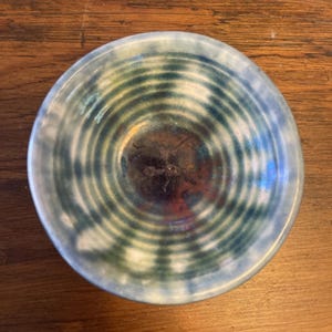 May include: A small, round ceramic bowl with concentric rings in blue, green, and white. The bowl's interior has a dark, mottled appearance, and the rim is pale blue. The bowl sits on a wooden surface.