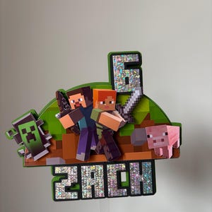 May include: A Minecraft cake topper with the characters Steve and Alex, a Creeper, and a pig. The number 6 is displayed with a sparkly finish, along with the name "ZACH". The topper is designed with a green and brown pixelated landscape.