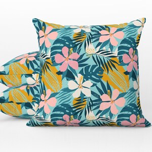 May include: Three square throw pillows with a tropical floral pattern. The design features pink and white hibiscus flowers, teal leaves, and yellow accents on a teal background. The pillows are arranged on a white background.