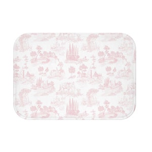 May include: A white bath mat with a pink toile pattern featuring repeating images of trees, houses, and a river.
