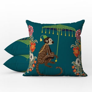 May include: Two teal throw pillows with a whimsical monkey design. The monkey is brown and holding a green umbrella. Floral accents in red, white, and yellow surround the monkey. The pillows are a decorative home accent.