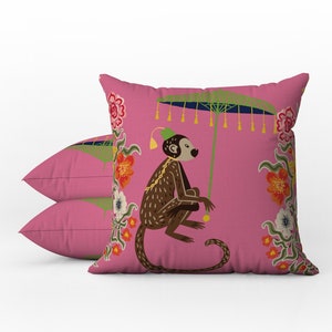 May include: Two pink throw pillows with a whimsical monkey design. The monkey is brown with a green hat and is holding a green parasol. The pillows have a floral border with red, orange, and white flowers.