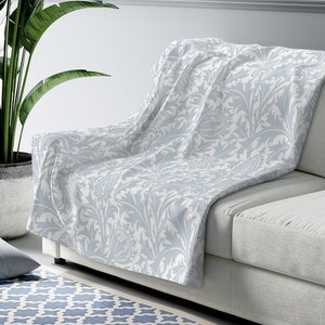 Könnte beinhalten: Eine hellblaue Wolldecke mit einem weißen Blumenmuster. Die Decke liegt über einem hellgrauen Sofa mit einem Kissen auf der Sitzfläche.