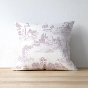 May include: Square decorative pillow with a lavender toile pattern depicting a classical landscape scene. The pillow is resting on a wooden surface against a white wall. The pillow features a detailed print of buildings, trees, and figures.