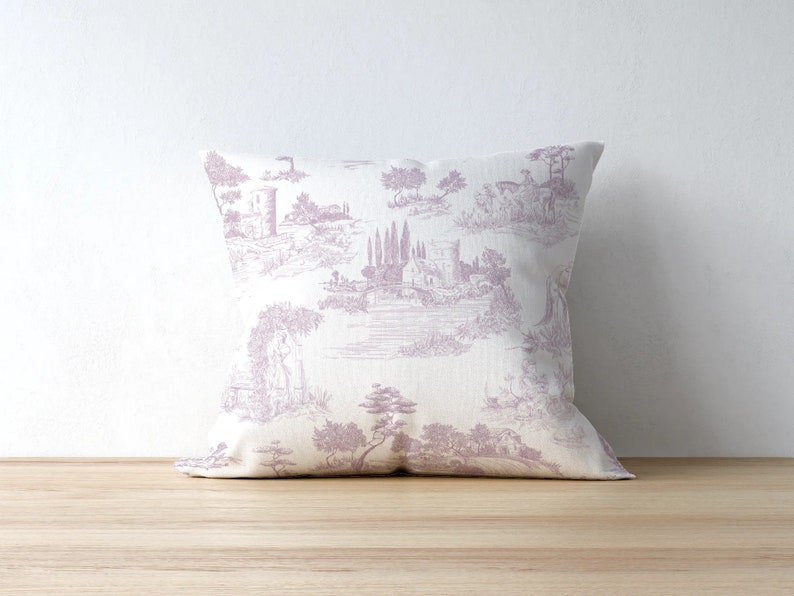 May include: Square decorative pillow with a lavender toile pattern depicting a classical landscape scene. The pillow is resting on a wooden surface against a white wall. The pillow features a detailed print of buildings, trees, and figures.