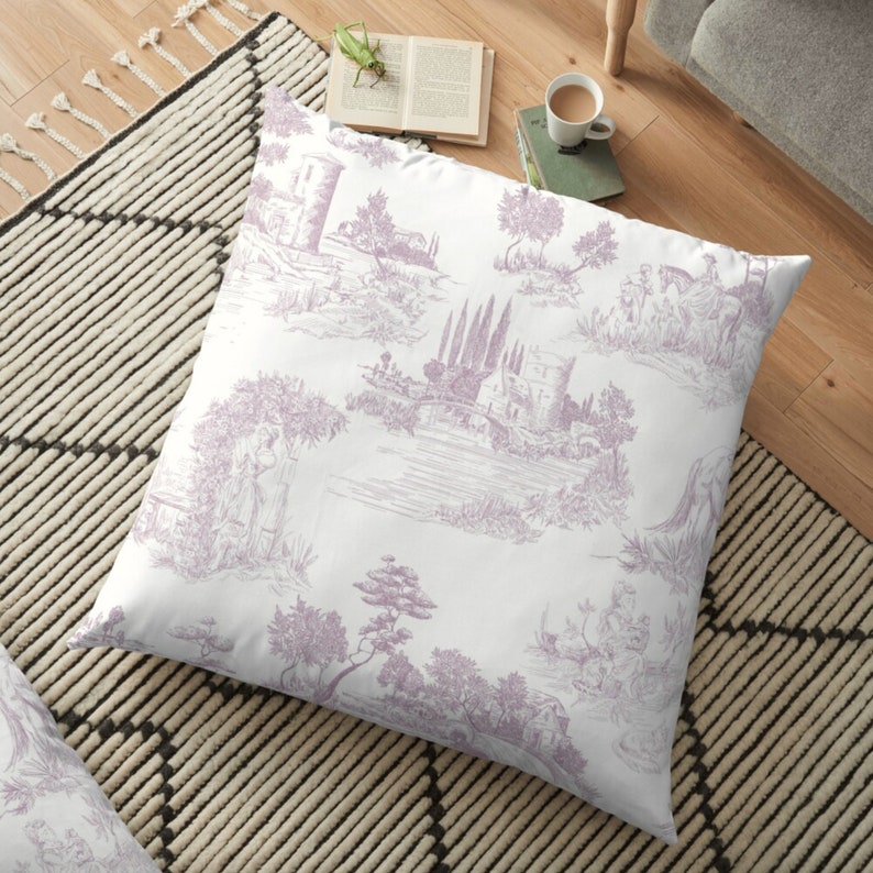 May include: A square floor pillow with a white background and a lavender toile pattern. The pattern depicts scenes of buildings, trees, and figures. The pillow is placed on a black and white patterned rug.