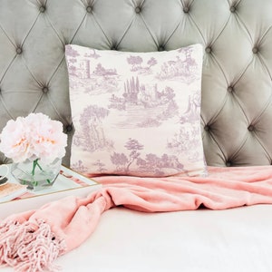 May include: A decorative pillow with a lavender and white toile design featuring a landscape scene. The pillow is placed against a gray, button-tufted headboard. A pink throw blanket, a tray with a cup, flowers, and a notebook complete the scene.
