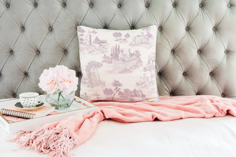 May include: A decorative pillow with a lavender and white toile design featuring a landscape scene. The pillow is placed against a gray, button-tufted headboard. A pink throw blanket, a tray with a cup, flowers, and a notebook complete the scene.