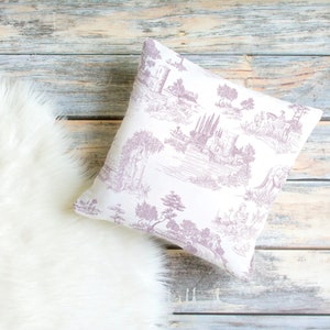 May include: Square throw pillow with a white background and a lavender toile pattern depicting a landscape scene. The pillow is resting on a light-colored wooden surface next to a white faux fur rug.