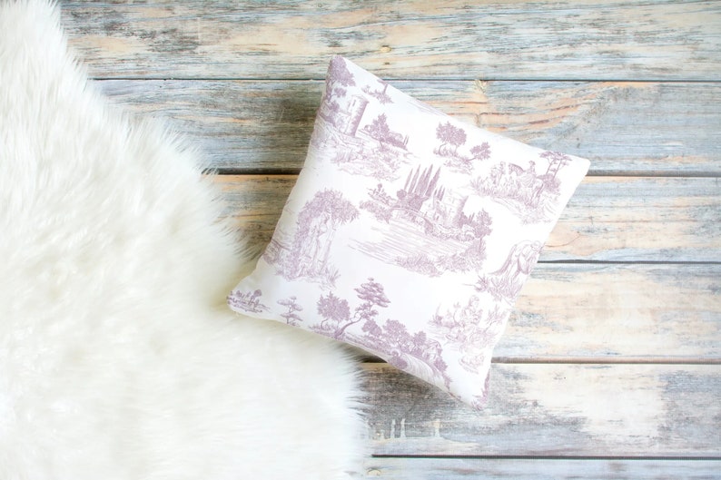May include: Square throw pillow with a white background and a lavender toile pattern depicting a landscape scene. The pillow is resting on a light-colored wooden surface next to a white faux fur rug.