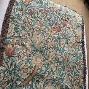 May include: A woven throw blanket with a floral pattern in shades of beige, teal, and rust. The design features stylized flowers and leaves. The blanket has a fringed edge.