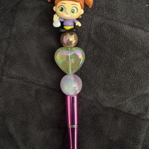 May include: A pink pen with a clear iridescent heart-shaped bead, a brown bead, and a small plastic figurine of a girl with brown hair and a purple shirt on top.