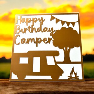 K&ouml;nnte beinhalten: Eine wei&szlig;e Karte mit goldenen Buchstaben, auf der "Happy Birthday Camper" steht. Die Karte hat ein ausgeschnittenes Design eines Wohnmobils, eines Baumes und eines Lagerfeuers.