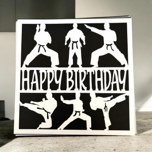 K&ouml;nnte beinhalten: Eine schwarz-wei&szlig;e Geburtstagskarte mit einer Silhouette von f&uuml;nf Personen in Karate-Posen. Die Karte sagt "Happy Birthday" in wei&szlig;en Buchstaben.