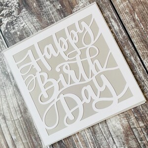 K&ouml;nnte beinhalten: Eine wei&szlig;e Karte mit grauem Hintergrund und den Worten "Happy Birthday" in einer dekorativen Schrift ausgeschnitten.
