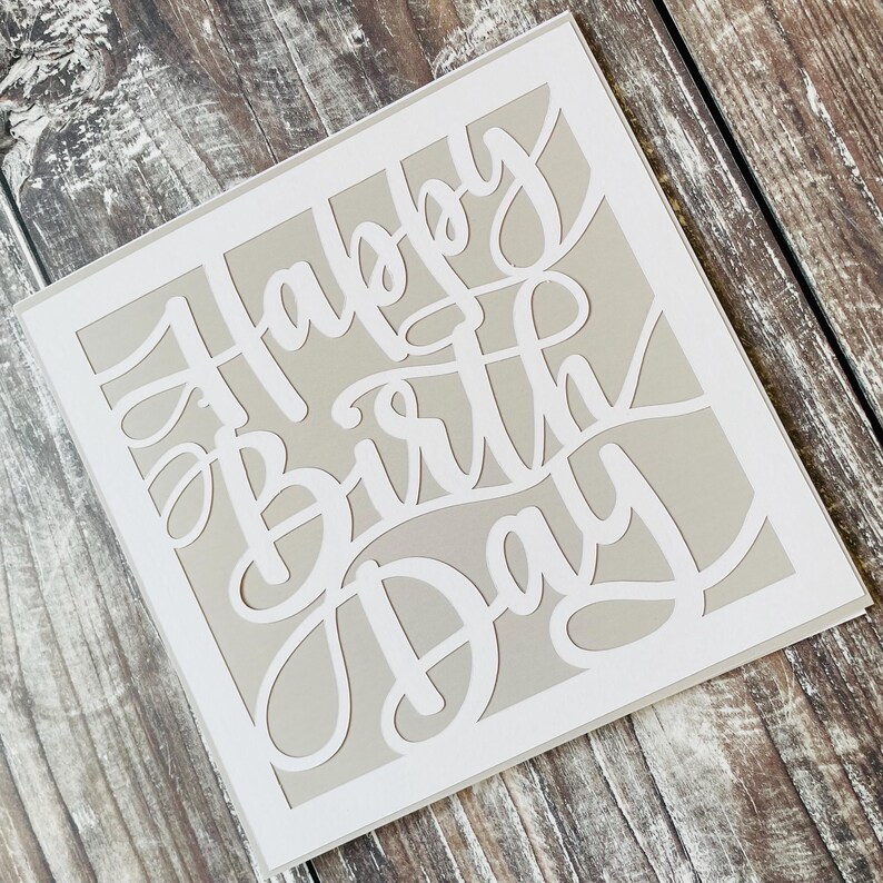 K&ouml;nnte beinhalten: Eine wei&szlig;e Karte mit grauem Hintergrund und den Worten "Happy Birthday" in einer dekorativen Schrift ausgeschnitten.