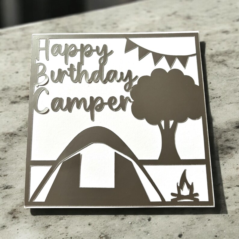 K&ouml;nnte beinhalten: Eine silberne Karte mit dem Text "Happy Birthday Camper" und einer Silhouette eines Zeltes, eines Baumes und eines Lagerfeuers.