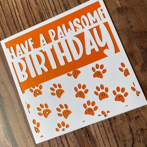 K&ouml;nnte beinhalten: Orangefarbene und wei&szlig;e Geburtstagskarte mit dem Text "Have a PAWsome Birthday" in wei&szlig;en Buchstaben. Die Karte hat ein ausgeschnittenes Design mit Pfotenabdr&uuml;cken.