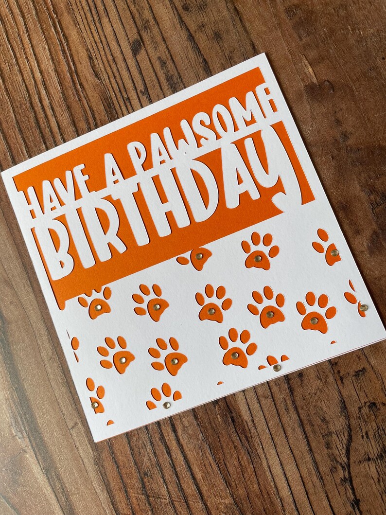 K&ouml;nnte beinhalten: Orangefarbene und wei&szlig;e Geburtstagskarte mit dem Text "Have a PAWsome Birthday" in wei&szlig;en Buchstaben. Die Karte hat ein ausgeschnittenes Design mit Pfotenabdr&uuml;cken.