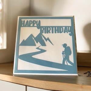 K&ouml;nnte beinhalten: Eine blaue Papierkarte mit einem ausgeschnittenen Design von Bergen, einem Fluss und einem Wanderer. Die Karte sagt "Happy Birthday" in blauer Schrift.