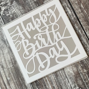 May include: A white card with a grey background featuring the words "Happy Birthday" cut out in a decorative font.