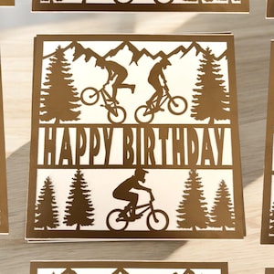 K&ouml;nnte beinhalten: Eine braune Karte mit einem ausgeschnittenen Design von Mountainbikern und B&auml;umen. Die Karte sagt "Happy Birthday" in fetter Schrift.