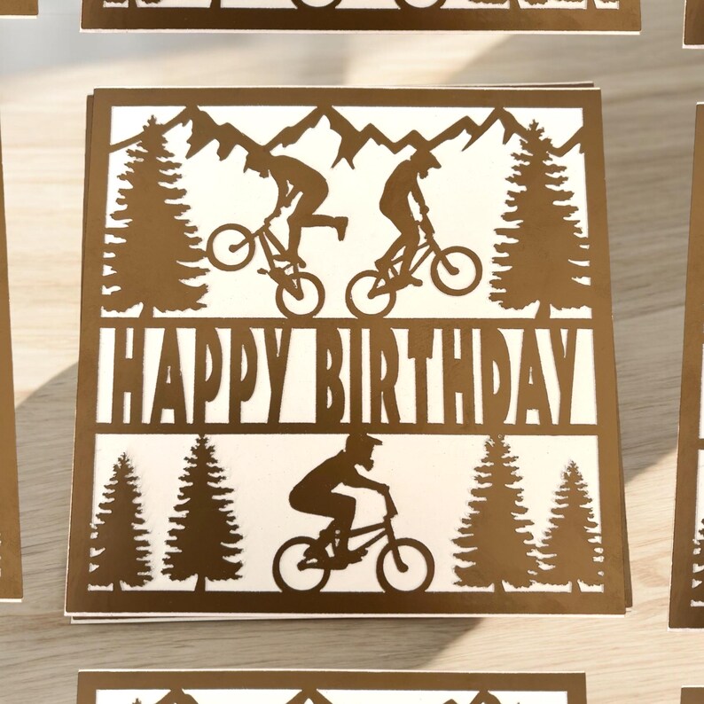 K&ouml;nnte beinhalten: Eine braune Karte mit einem ausgeschnittenen Design von Mountainbikern und B&auml;umen. Die Karte sagt "Happy Birthday" in fetter Schrift.
