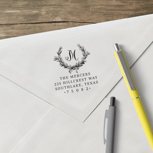 May include: A white envelope with a black return address stamp. The stamp features a wreath with the letter "M" in the center. The text on the stamp reads "The Mercers, 235 Hillcrest Way, Southlake, Texas, 75082."