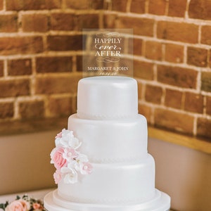 May include: A white wedding cake with pink and white flowers on the bottom tier. A clear acrylic topper with the text "Happily ever after" and the names "Margaret & John" and the date "10.14.20" is on top of the cake.