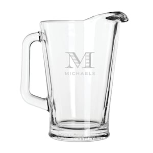 May include: A clear glass pitcher with a handle and a spout. The pitcher is engraved with the letter "M" and the name "Michaels".
