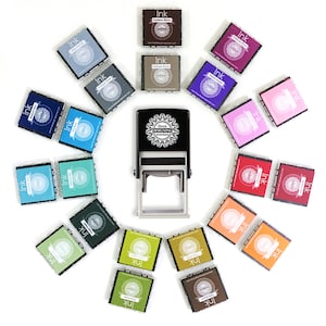 May include: A black self-inking stamp with the text "three designing women" and a circular design. The stamp is surrounded by 16 small ink pads in various colors, including blue, green, red, pink, purple, brown, and gray.
