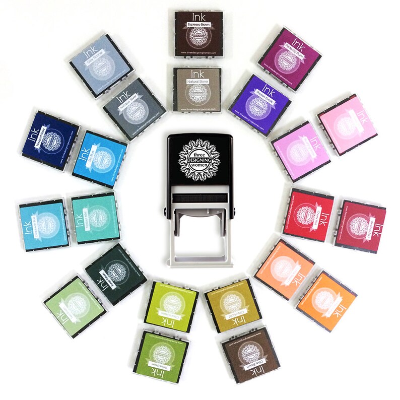 May include: A black self-inking stamp with the text "three designing women" and a circular design. The stamp is surrounded by 16 small ink pads in various colors, including blue, green, red, pink, purple, brown, and gray.