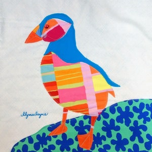 May include: A colorful, patchwork fabric bird with a blue head and red eye, standing on a green and blue floral patterned fabric background. The bird is made of different colored fabric scraps, including pink, orange, yellow, and red. The text "lilymaylayne" is embroidered in blue thread on the fabric background.