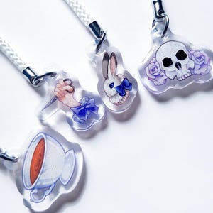 Peut inclure: Charmes en acrylique transparents avec des cordons blancs. Les breloques présentent des illustrations d'une main tenant un couteau, d'un lapin avec un nœud bleu, d'un crâne avec des roses violettes et d'une tasse de thé.