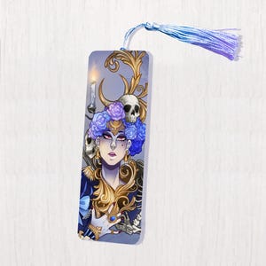 Peut inclure: Un marque-page représentant une femme portant une couronne de fleurs bleues et violettes, entourée de crânes et d'une bougie. L'arrière-plan est gris clair avec des accents dorés.