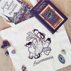Può includere: Una borsa di tela bianca con un'illustrazione in bianco e nero di un pesce in una nuvola. Il testo "Oniromancy." è stampato sotto il pesce. La borsa è appoggiata su una superficie di legno con diversi piccoli cristalli viola e blu sparsi intorno.