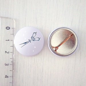 May include: A white button featuring a silver pair of scissors with an ornate design and sparkles on a white background. The reverse of the button is silver with a pin.